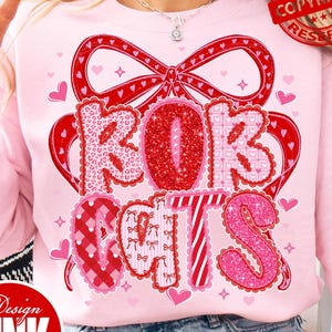 May include: A light pink sweatshirt with a red bow and the words "ROK CATS" in a decorative font. The letters are in various patterns and colors, including red glitter and pink gingham. Small hearts are scattered around the design.