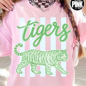 Peut inclure: T-shirt rose clair avec le mot "tigers" en vert et une illustration de tigre vert. Le t-shirt a des rayures blanches verticales. Un logo "Design Pink Art" est dans le coin supérieur droit.