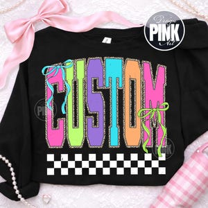 May include: Black sweatshirt with the word "CUSTOM" in large, colorful, block letters. The letters have a leopard print fill and are outlined in various colors. The design includes bows and a checkered border.