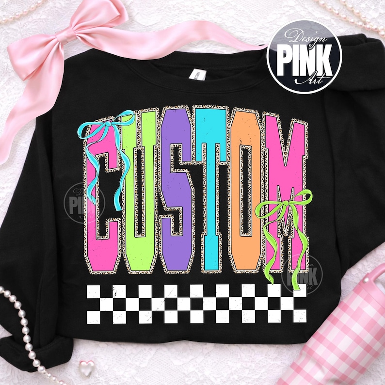 May include: Black sweatshirt with the word "CUSTOM" in large, colorful, block letters. The letters have a leopard print fill and are outlined in various colors. The design includes bows and a checkered border.