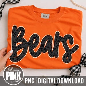 May include: A bright orange short-sleeved t-shirt featuring the word Bears in a large, cursive, black and grey leopard print font with a white outline. The inner neck label reads Comfort. A circular logo in the corner says Design Pink Art.