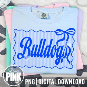 May include: Light blue t-shirt with the word "Bulldogs" in a blue script font, framed by a blue scalloped border and bow. The shirt is folded and stacked with other pastel-colored shirts. The text "PNG | DIGITAL DOWNLOAD" is at the bottom.