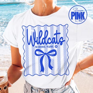 May include: White t-shirt with a blue and white design. The design features the word "Wildcats" in a script font, a blue bow, and vertical stripes with polka dots. The shirt also has the text "SCHOOL NAME".