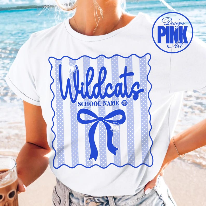 May include: White t-shirt with a blue and white design. The design features the word "Wildcats" in a script font, a blue bow, and vertical stripes with polka dots. The shirt also has the text "SCHOOL NAME".