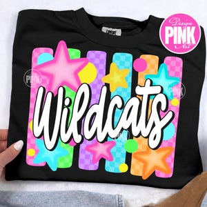 May include: Black sweatshirt with the word "Wildcats" in white script. The design features colorful stars and vertical stripes in pink, blue, orange, green, and purple. The design includes the text "Design PINK Art" in a pink circle.