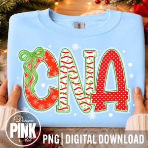 Pode incluir: Uma camisola azul-clara de gola redonda com as letras CNA num design festivo. O C é vermelho com flocos de neve brancos e um laço verde, o N tem riscas vermelhas e verdes, e o A é vermelho com bolas brancas, tudo com contorno branco.