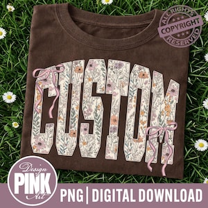 May include: A brown t-shirt featuring the word "CUSTOM" in large, floral letters. The letters are cream-coloured and filled with flowers in pink, orange, and purple. Pink ribbons are tied around the letters. The shirt is displayed on a bed of green grass.