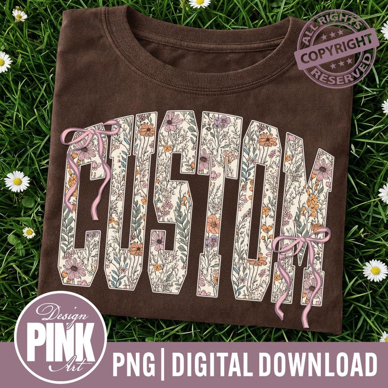 May include: A brown t-shirt featuring the word "CUSTOM" in large, floral letters. The letters are cream-coloured and filled with flowers in pink, orange, and purple. Pink ribbons are tied around the letters. The shirt is displayed on a bed of green grass.