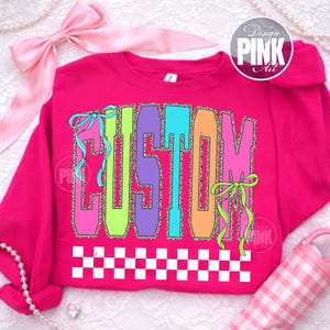 May include: A hot pink sweatshirt with the word "CUSTOM" in large, colorful letters. The letters are outlined in leopard print and have ribbons attached. A white and black checkered pattern is at the bottom.