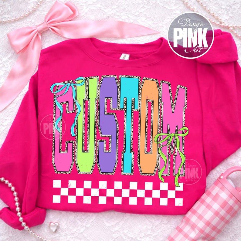 May include: A hot pink sweatshirt with the word "CUSTOM" in large, colorful letters. The letters are outlined in leopard print and have ribbons attached. A white and black checkered pattern is at the bottom.