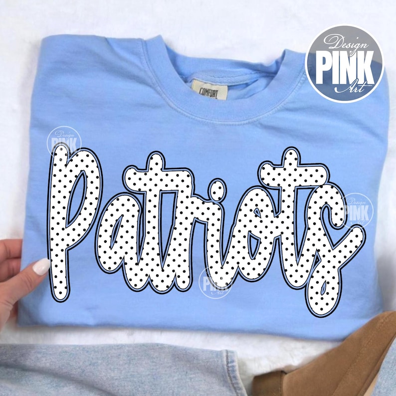 May include: Light blue sweatshirt with the word "Patriots" in a white, polka-dotted font. The word is outlined in black. The sweatshirt has a crew neck and a tag that says "Comfort". A "Design Pink Art" logo is in the upper right corner.