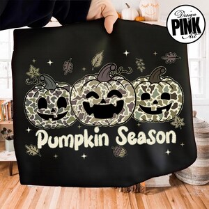 Puede incluir: Tela negra con un dise&ntilde;o que presenta tres calabazas sonrientes con un patr&oacute;n de camuflaje. Las calabazas est&aacute;n dispuestas horizontalmente y el texto "Pumpkin Season" est&aacute; debajo. Hojas y estrellas decorativas rodean las calabazas.