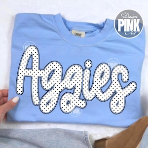 Peut inclure: Un sweat-shirt bleu clair avec le mot "Aggies" en blanc &agrave; pois. Le sweat-shirt porte une &eacute;tiquette "Comfort" et un logo circulaire "Design Pink Art".