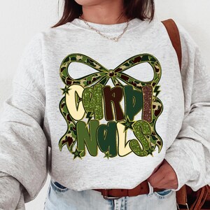 May include: A light gray sweatshirt featuring the word "CARDINALS" in a decorative font with a camouflage bow design. The letters are in shades of green, brown, and cream, with star accents.