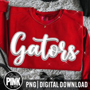May include: A bright red long-sleeve sweatshirt featuring the word Gators in large, white, cursive script lettering with a silver glitter outline. The garment has a crew neckline and a visible interior tag. The design is centered on the front.