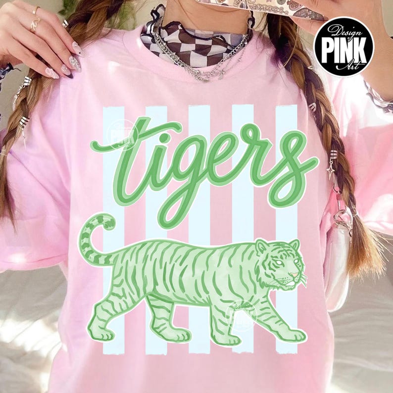 May include: Pink t-shirt with the word "tigers" in green script and a green tiger illustration. The shirt has vertical white stripes. A "Design Pink Art" logo is in the upper right corner.