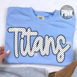May include: Light blue sweatshirt with the word "Titans" in large, white, polka-dotted letters. The word is outlined in black. A "Design Pink Art" logo is in the upper right corner.