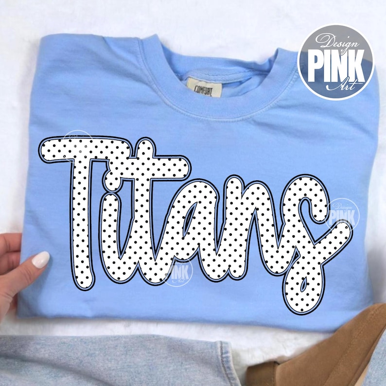 May include: Light blue sweatshirt with the word "Titans" in large, white, polka-dotted letters. The word is outlined in black. A "Design Pink Art" logo is in the upper right corner.
