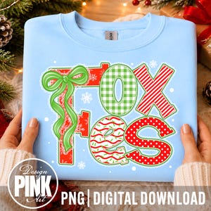 May include: A light blue crewneck sweatshirt featuring a large graphic that spells out the word FOXES. The letters are decorated with green gingham, red and white stripes, and red polka dots, accented by a large green ribbon and snowflakes.