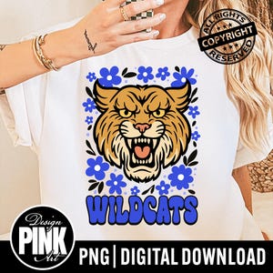 Puede incluir: Camiseta blanca con un dise&ntilde;o de gato mont&eacute;s marr&oacute;n y beige con la boca abierta, rodeado de flores azules. La palabra "WILDCATS" es en azul, con el texto "Design PINK Art" y "PNG | DIGITAL DOWNLOAD".