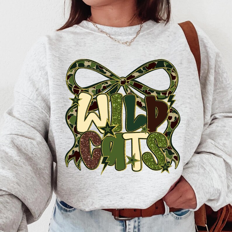 May include: A light gray sweatshirt with the words "WILD CATS" in a camouflage and glitter design. The text is framed by a camouflage bow. The sweatshirt has long sleeves and a crew neck.