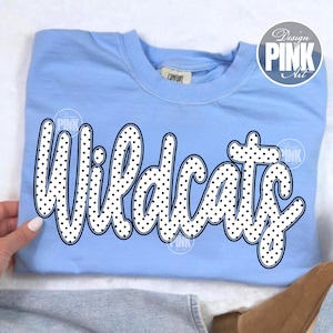 May include: A light blue sweatshirt with the word "Wildcats" in large, white, polka-dotted letters. The sweatshirt has a crew neck and long sleeves. The text is outlined in black.