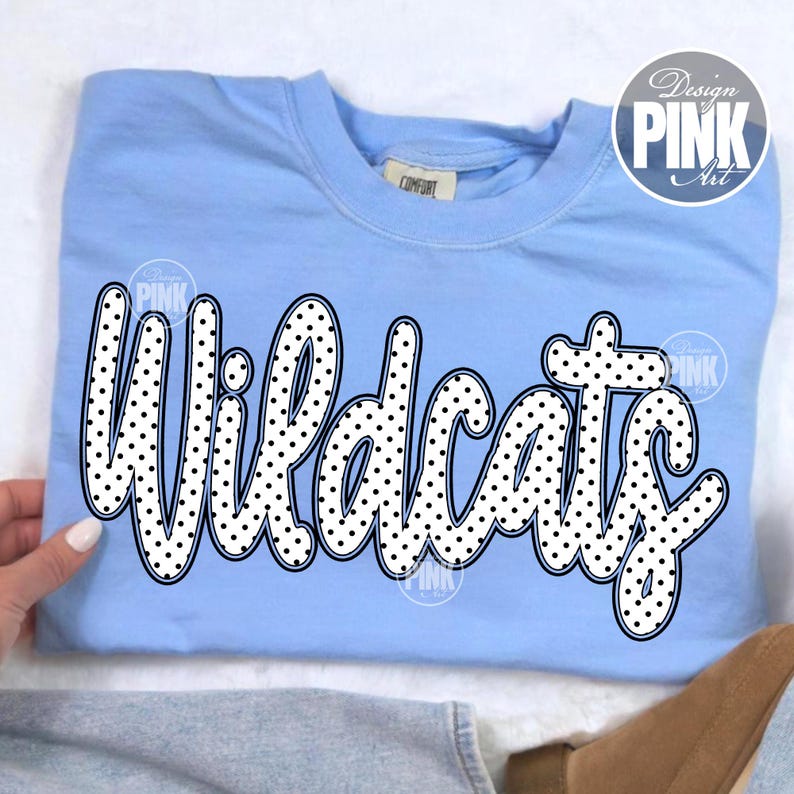 May include: A light blue sweatshirt with the word "Wildcats" in large, white, polka-dotted letters. The sweatshirt has a crew neck and long sleeves. The text is outlined in black.
