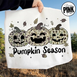 Puede incluir: Tela blanca con un dise&ntilde;o de "Pumpkin Season". El dise&ntilde;o presenta tres calabazas sonrientes con patrones de camuflaje en tonos verdes y marrones. Hojas y estrellas rodean las calabazas. Las palabras "Design Pink Art" est&aacute;n en un c&iacute;rculo en la esquina superior derecha.