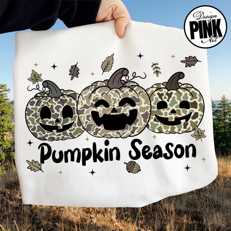Puede incluir: Tela blanca con un dise&ntilde;o de "Pumpkin Season". El dise&ntilde;o presenta tres calabazas sonrientes con patrones de camuflaje en tonos verdes y marrones. Hojas y estrellas rodean las calabazas. Las palabras "Design Pink Art" est&aacute;n en un c&iacute;rculo en la esquina superior derecha.