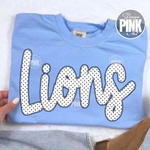 Może przedstawiać: Jasnoniebieska bluza z napisem "Lions" dużymi, białymi literami w kropki. Bluza ma okrągły dekolt i długie rękawy. Słowa "Design Pink Art" znajdują się w okręgu w prawym g&oacute;rnym rogu.