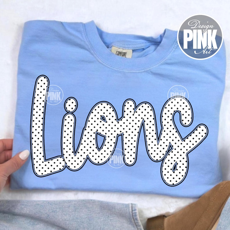 Może przedstawiać: Jasnoniebieska bluza z napisem "Lions" dużymi, białymi literami w kropki. Bluza ma okrągły dekolt i długie rękawy. Słowa "Design Pink Art" znajdują się w okręgu w prawym g&oacute;rnym rogu.