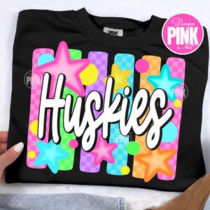 Pu&ograve; includere: Felpa nera con la scritta "Huskies" in corsivo bianco. Il design include stelle colorate e barre verticali rosa, verdi, arancioni e viola, con un motivo a scacchi. La felpa ha un girocollo.