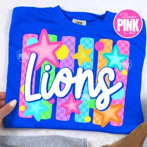 May include: Royal blue sweatshirt with the word "Lions" in white script. The design includes colorful stars and vertical stripes in pink, green, orange, and purple. The sweatshirt has a crew neck and a small tag that says "Comfort".