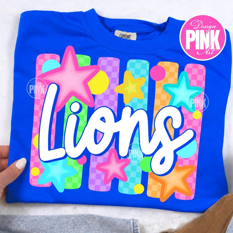 May include: Royal blue sweatshirt with the word "Lions" in white script. The design includes colorful stars and vertical stripes in pink, green, orange, and purple. The sweatshirt has a crew neck and a small tag that says "Comfort".