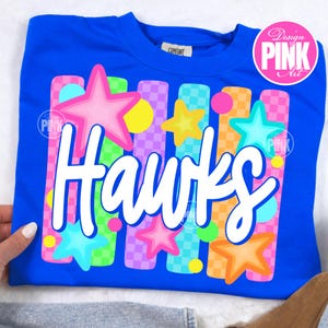 May include: A royal blue sweatshirt with the word "Hawks" in white script. The design includes colorful stars, circles, and vertical stripes in pink, yellow, green, orange, and purple. The shirt has a "Comfort" tag.