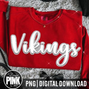 May include: A bright red long-sleeve crewneck sweatshirt featuring the word Vikings in a large, white, three-dimensional script font with a silver glitter outline. The garment has a ribbed neckline and a visible interior brand label.