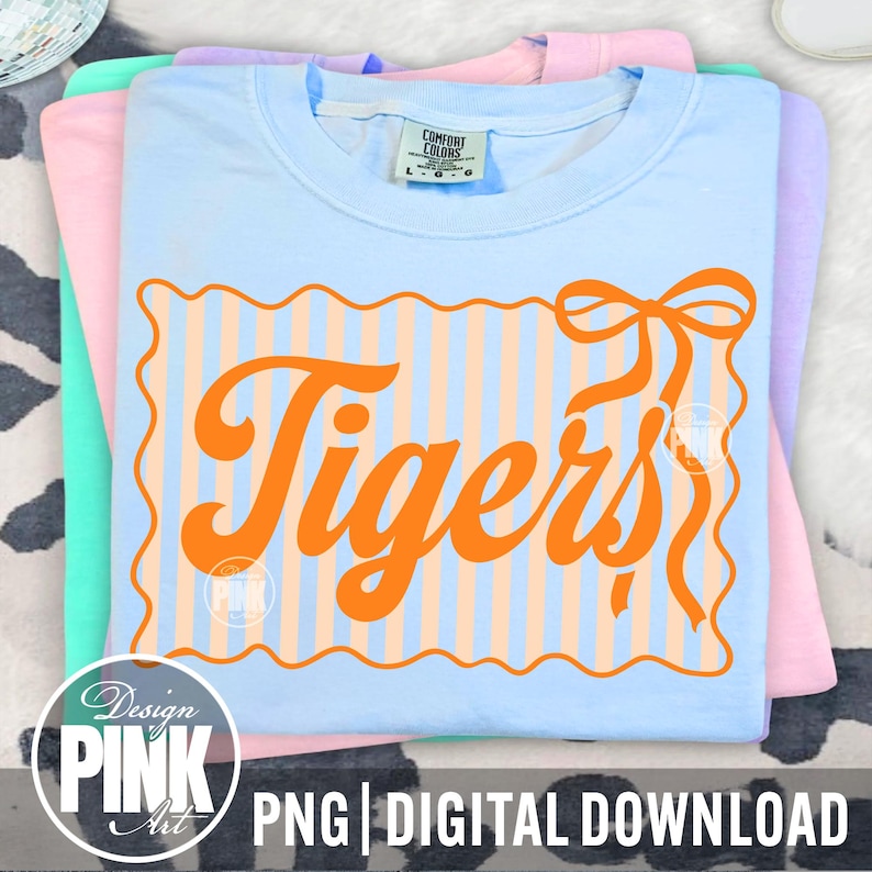 May include: A stack of Comfort Colors t-shirts in pastel colors: light blue, pink, and green. The top shirt is light blue with an orange design that says "Tigers" in a retro font. The design includes a striped background and a bow.