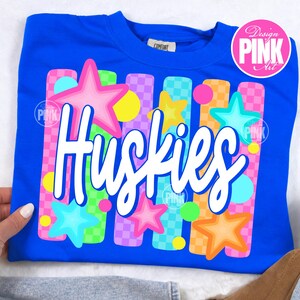 Pu&ograve; includere: Una felpa blu reale con la scritta "Huskies" in corsivo bianco. Il design include stelle colorate e strisce verticali rosa, arancioni, verdi e viola, con un logo circolare rosa con la scritta "Design PINK Art".