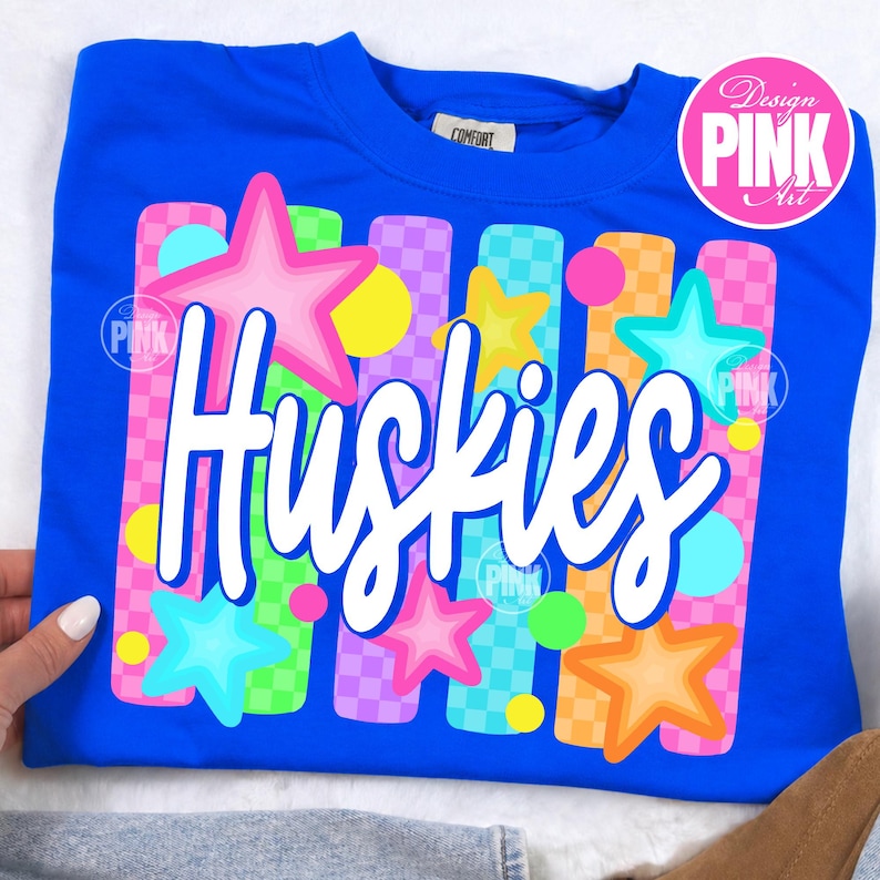 Pu&ograve; includere: Una felpa blu reale con la scritta "Huskies" in corsivo bianco. Il design include stelle colorate e strisce verticali rosa, arancioni, verdi e viola, con un logo circolare rosa con la scritta "Design PINK Art".