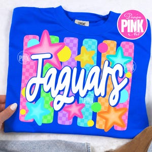 May include: A royal blue sweatshirt with the word "Jaguars" in white script. The design includes colorful stars, circles, and vertical stripes in pink, yellow, orange, green, and purple. The shirt has a crew neck and a tag that says "Comfort".