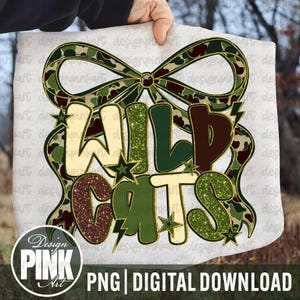 May include: A white background with the words "WILD CATS" in a decorative font, with a camouflage bow and stars. The text is in shades of green, brown, and gold. The image also includes the text "PNG | DIGITAL DOWNLOAD" and a logo for "Design PINK Art".