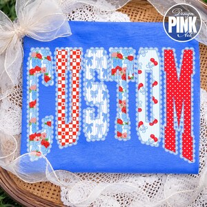 May include: A blue t-shirt featuring the word "CUSTOM" in a decorative font. The letters are filled with patterns including red gingham, blue bows, red roses, and cherries. The design is framed with a scalloped edge.