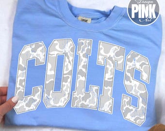 May include: A light blue crewneck t-shirt featuring the word COLTS in large, block-style letters across the chest. The letters have a white and grey camouflage pattern with a distressed, weathered texture. A small tag is visible at the neckline.