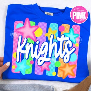 May include: A royal blue crew neck sweatshirt with the word "Knights" in white script. The design includes colorful stars, circles, and vertical stripes in pink, yellow, green, and orange. The sweatshirt has a "Design Pink Art" logo.