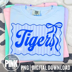 May include: A stack of pastel-colored t-shirts. The top shirt is light blue with a blue striped design and the word "Tigers" in a bold, cursive font. The design includes a bow. The bottom of the image has the text "PNG | DIGITAL DOWNLOAD".