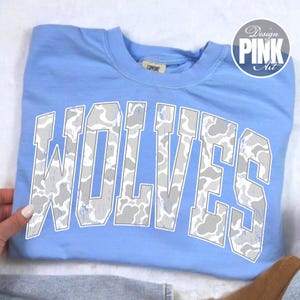 May include: A light blue crew neck t-shirt featuring the word WOLVES in large, block-style letters across the chest. The letters have a white and grey camouflage pattern with a distressed texture. A circular logo in the top right reads Design PINK Art.