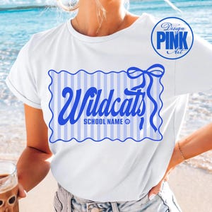 May include: White t-shirt with a blue and white striped design featuring the word "Wildcats" in blue script. The shirt includes a blue bow and the text "SCHOOL NAME" below the word "Wildcats". A "Design Pink Art" logo is in the upper right corner.