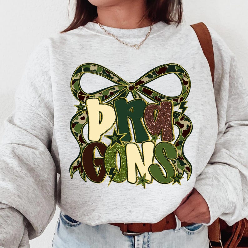 May include: A light gray sweatshirt with the word "DRAGONS" in a decorative design. The letters are in various colors, including tan, green, and brown, with a camouflage bow and star accents.