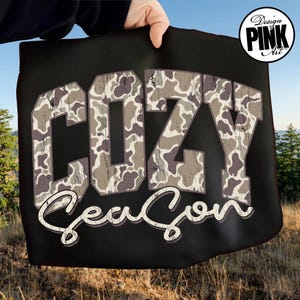 May include: Black tote bag with the words "COZY Season" in a camouflage pattern and white script. The bag is held up by a hand, with a "Design Pink Art" logo in the upper right corner. The background shows a natural outdoor scene.