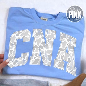 May include: A light blue crewneck t-shirt featuring the letters CNA in a large, arched collegiate font. The letters are filled with a white and gray cow print pattern. The shirt has a visible brand tag inside the collar area.